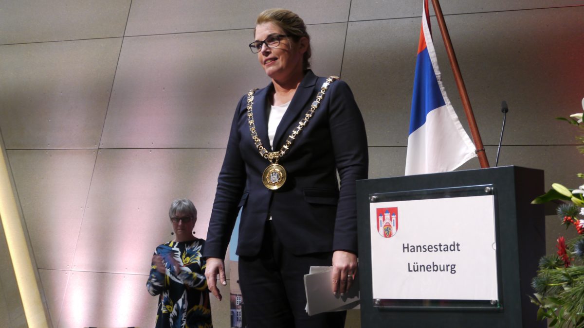 Claudia Kalisch steht neben einem Pult mit der Aufschrift "Hansestadt Lüneburg". Sie trägt eine goldene Kette, die ihr als neue OB überreicht wurde