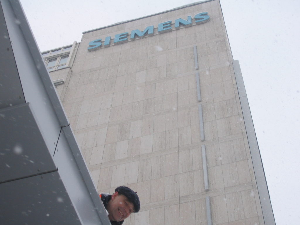 Karsten guck von oben, er guckt vin einer Kante eines Vordach heraus. Oben ganz oben auf dem Gebäude, das Siemens Logo. Es schneit, Karsten lächelt