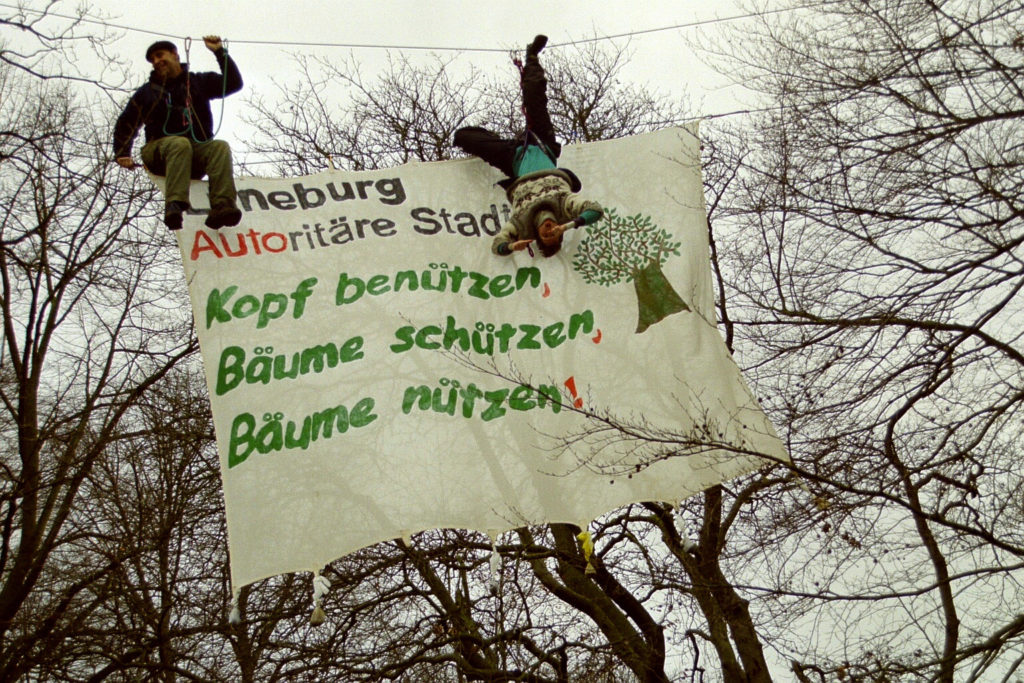 Zwei Personen in einer ZTraverse (Seil) mit Banner: Lüneurg Autoritäre Stadt Kopf benutzen Bäume schützen Bäume nützen! und ein Baum als Bild