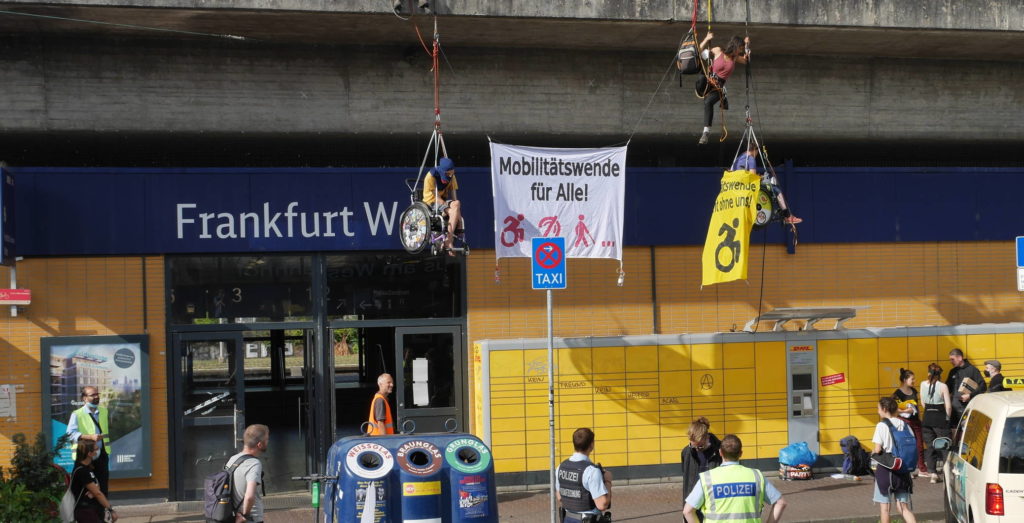 zwei kletter*innen sitzen in Seilen in ihrem Rollstuhl an einer Gebäudefassade, zwischen ihnen eine Banner "Mobilitätswende für alle" Eine Person hat ein Handbanner "Mobilitätswende nicht ohne uns" und zwei Polizisten gucken nach oben