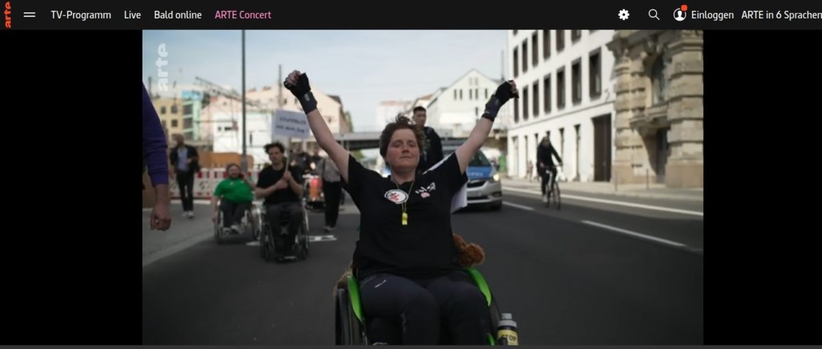 screenshot Arte Beitrag, ich fahre Rollstuhl und hebe die Arme als wäre ich frei und trage ISL t-Shirt