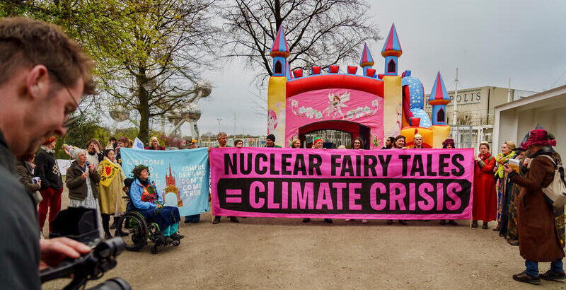 Menschen als Märchenfiguren gekleidet, eine Schloss-Hüpfburg und großes rosa Banner Nuclear fary tale = climate crisis
