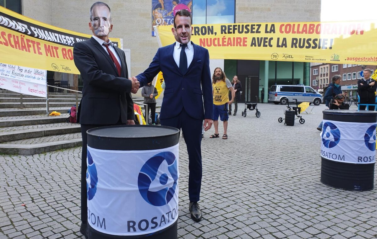 2 Menschen im Anzug mit Masken von Putin und Macron gegben sich die Hand, Drumherum Fässer mit dem Logo Rosatom und Banner gegen die Atomgeschäfte Macron Putin
