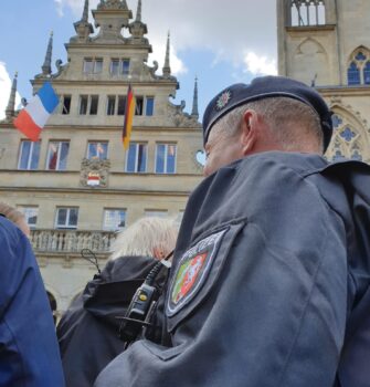 mein Blick, im Rollstuhlsitzend, ein großer Polizist vor mir, menschenmenge und hinten das Rathaus mit Macron am Balkon der winkt mit seiner Frau