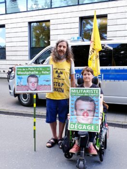 Zwei Menschen mit Plakaten mit Macrons Gesicht mit roter nase und Spruch auf Französisch "militariste dégage" und  "prix de la paix hihi la Westfalie est folle"